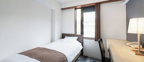 Une chambre d’hôtel avec un lit simple, un bureau avec une lampe et une fenêtre avec des rideaux.