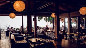 An outdoor seating area with wooden tables and sofas, large windows offering views of the sea, and hanging paper lanterns. - 15 Palms Beach Resort (Ko Chang)