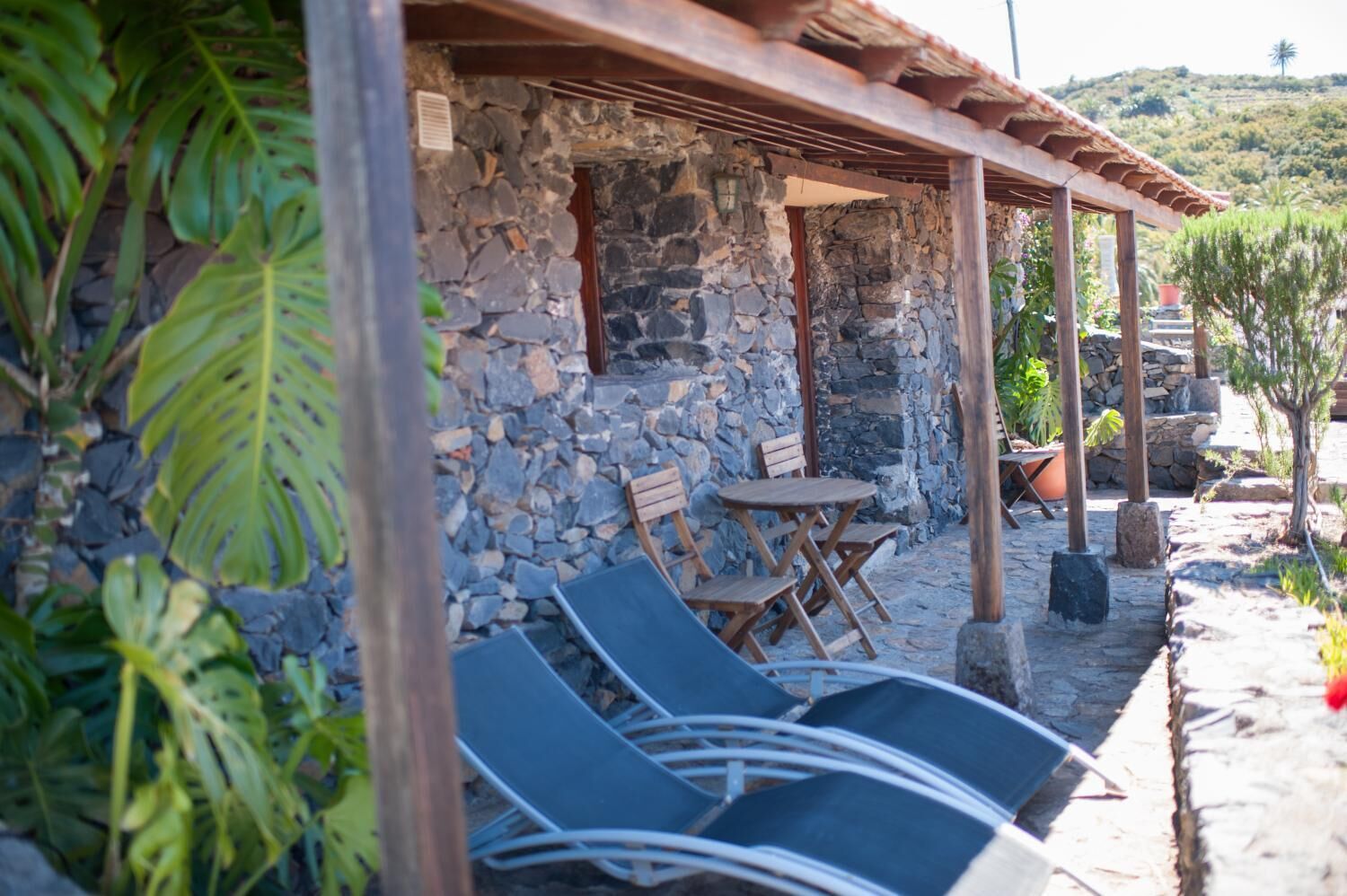 Un edificio de piedra con una pérgola de madera, asientos al aire libre y plantas en macetas.