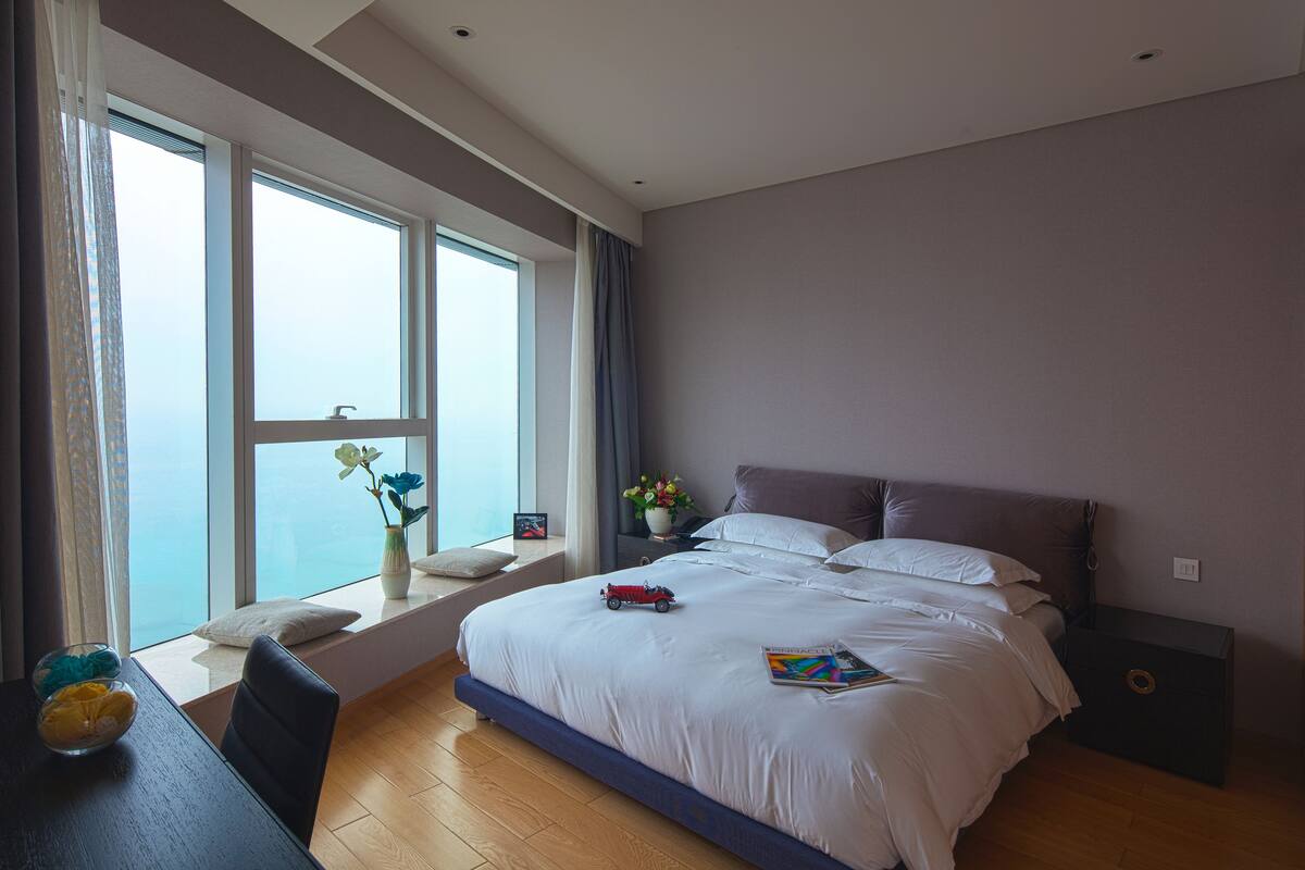 Une chambre à coucher moderne avec un grand lit, une table de chevet, une fenêtre avec des rideaux et une vue sur le ciel.