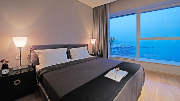 Een moderne hotelkamer met een groot bed, twee bedlampjes, een nachtkastje met een klok, en uitzicht op de stad door een groot raam.