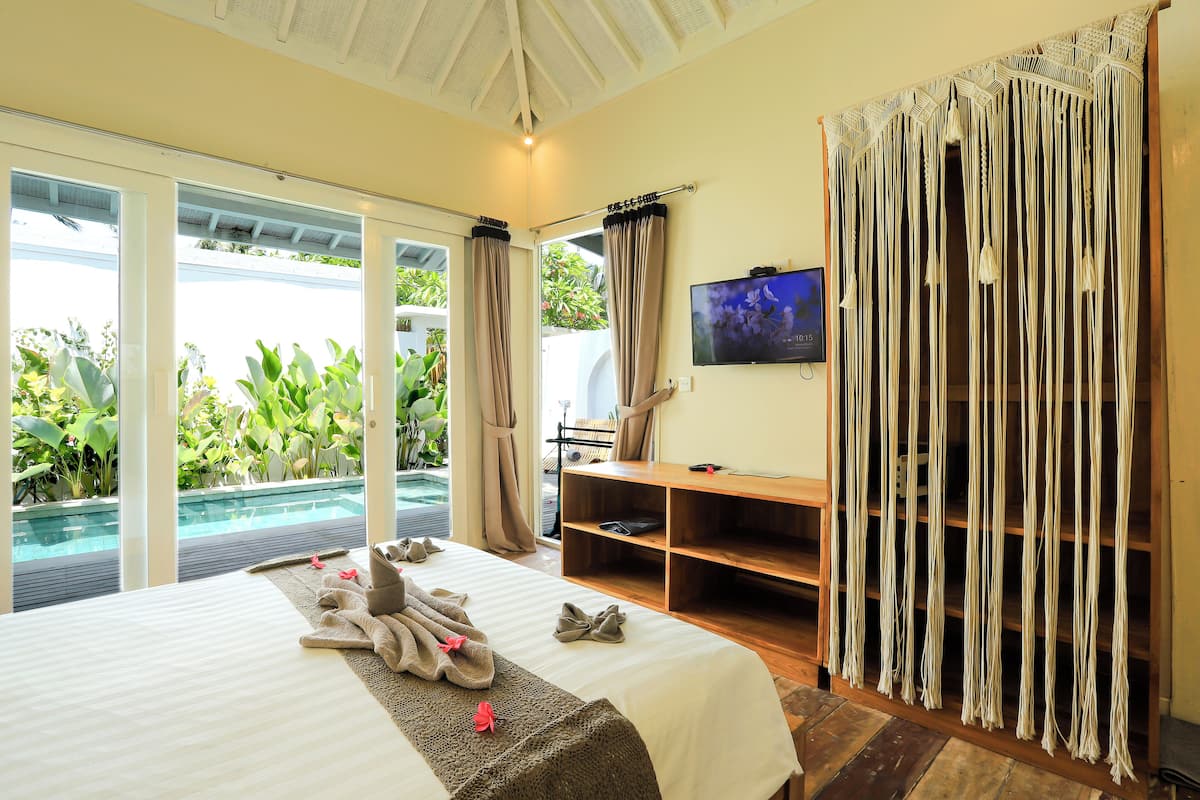 A hotel room with a bed, a TV, a wooden shelf, and a view of a pool and greenery.