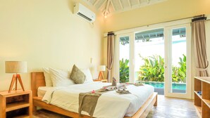 A bedroom with a wooden bed, a bedside table with a lamp, a window with curtains, and a view of greenery outside. - Villas Edenia (Gili Trawangan)