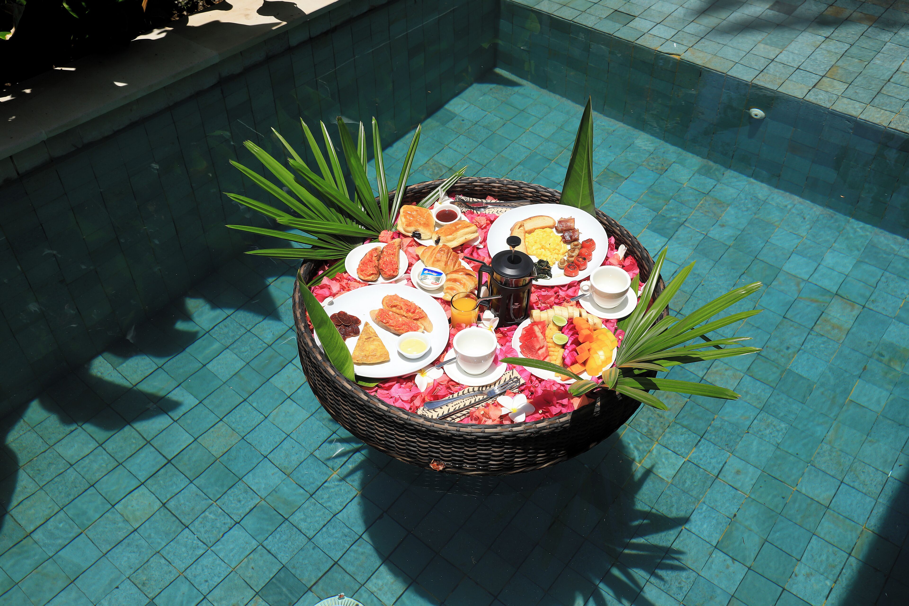Un panier en osier rempli de divers aliments pour le petit-déjeuner, posé sur une terrasse carrelée près de la piscine.
