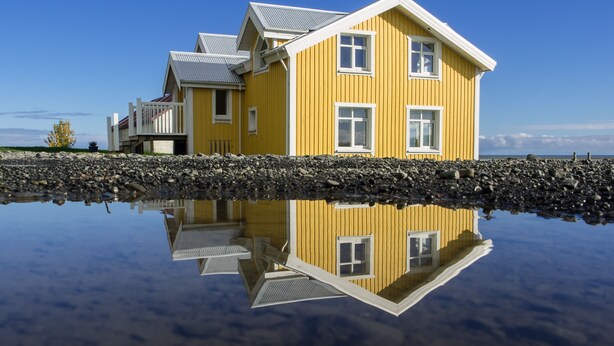 A yellow house with a balcony, reflecting in a calm body of water.