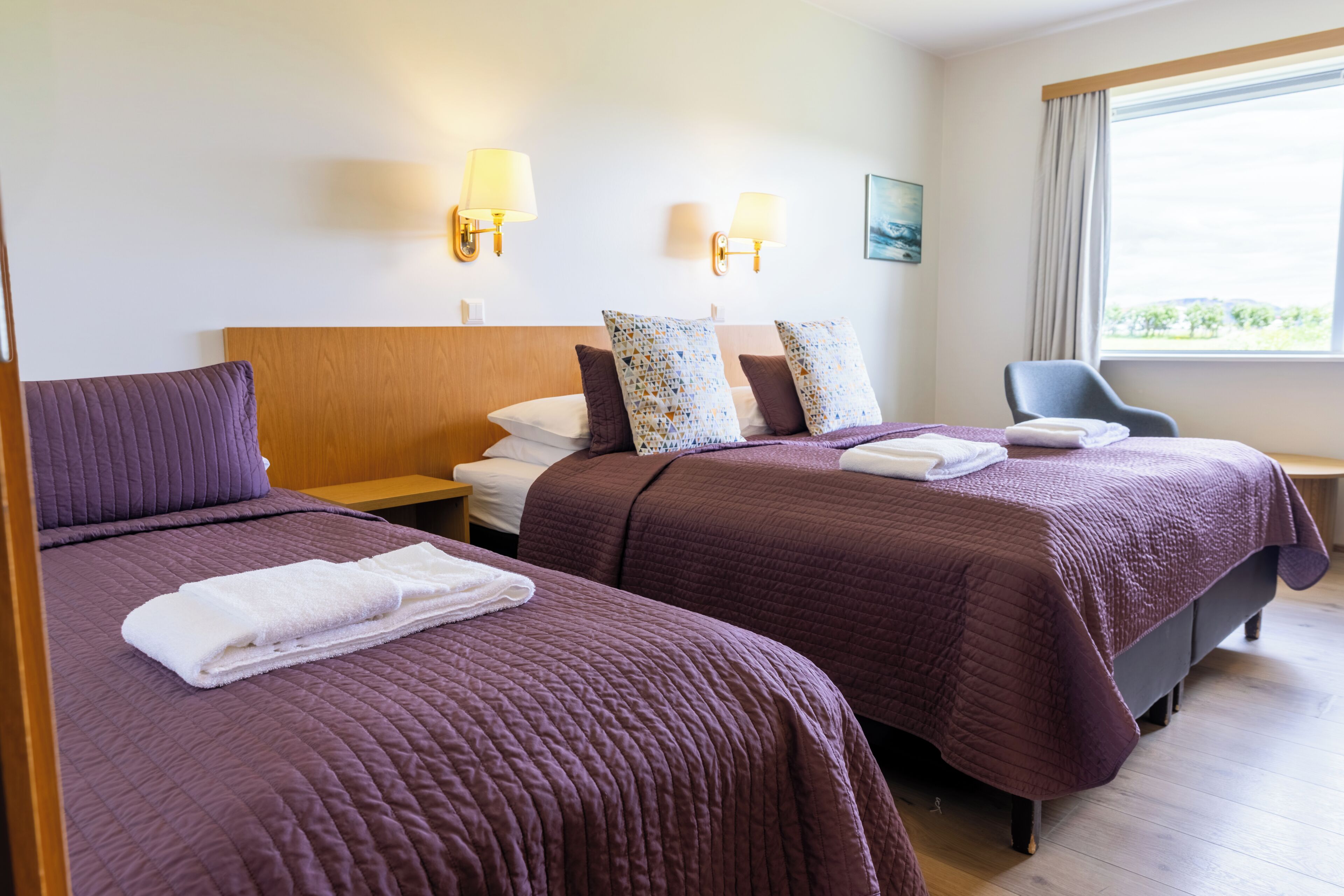 A hotel room with two beds, a desk, and a chair. There are wall-mounted lamps and a painting on the wall.