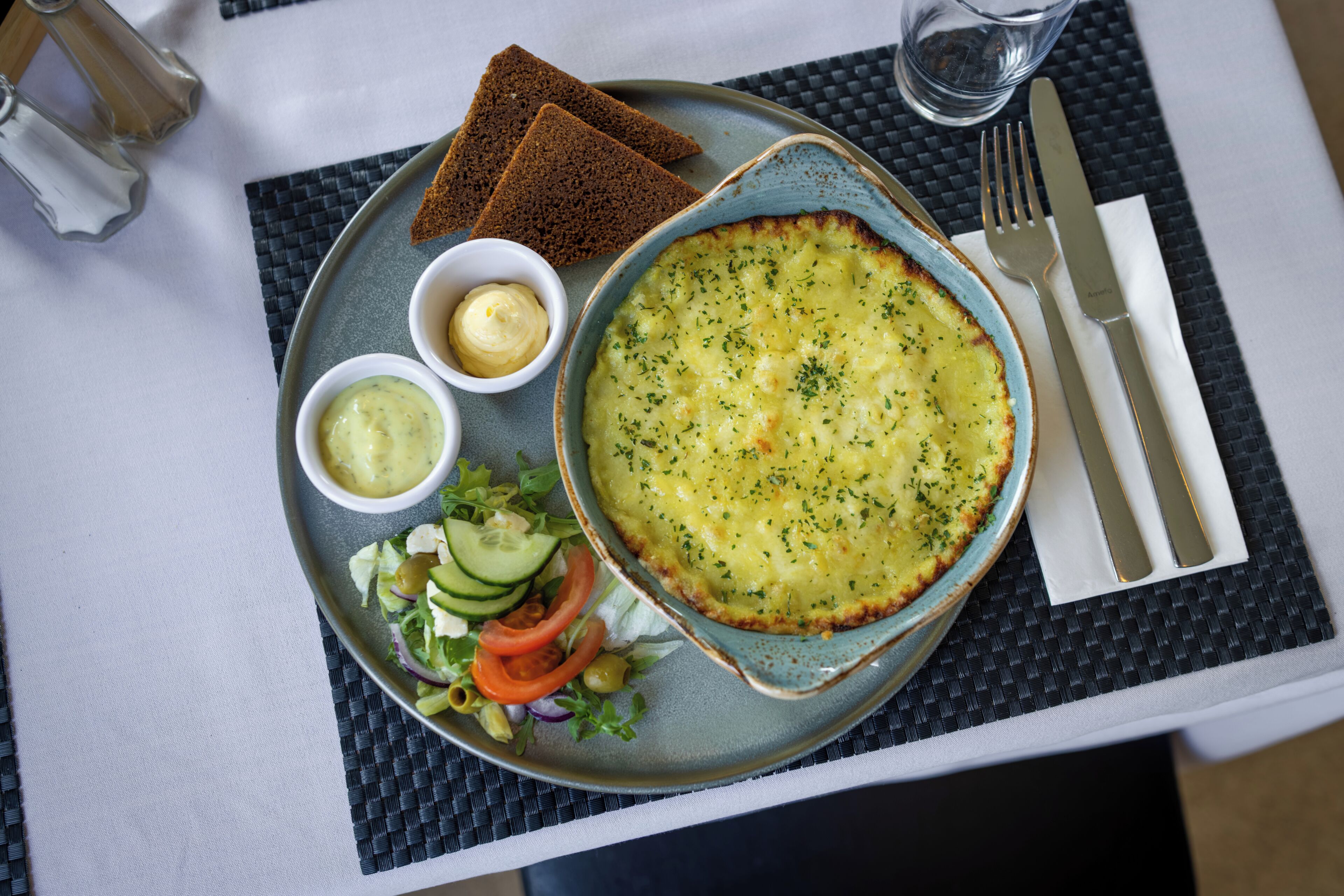 A plate with a baked dish, two small bowls of sauce, a salad, and slices of bread on a table.