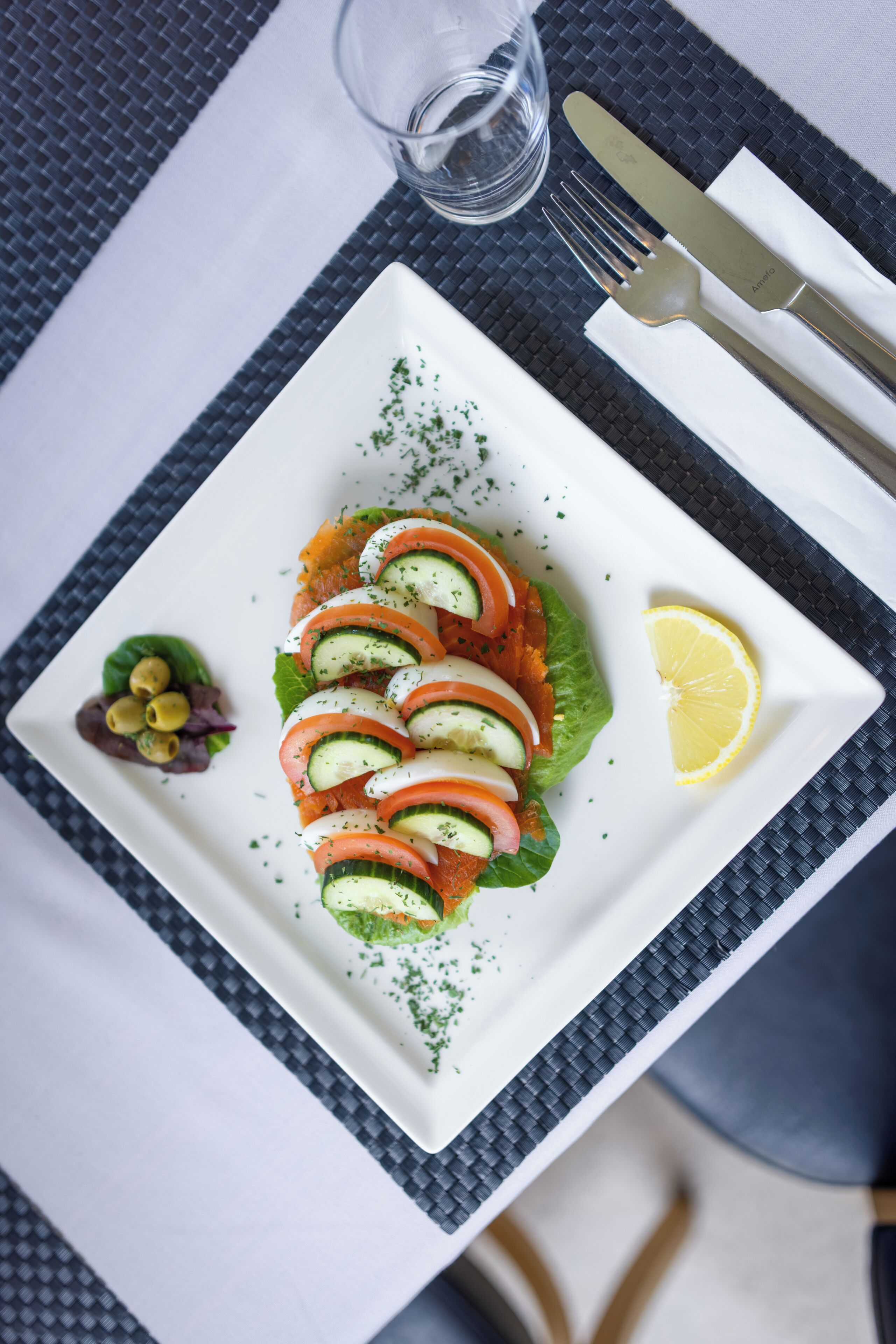 Un plato cuadrado blanco con ensalada, un gajo de limón y un recipiente pequeño de aceitunas.