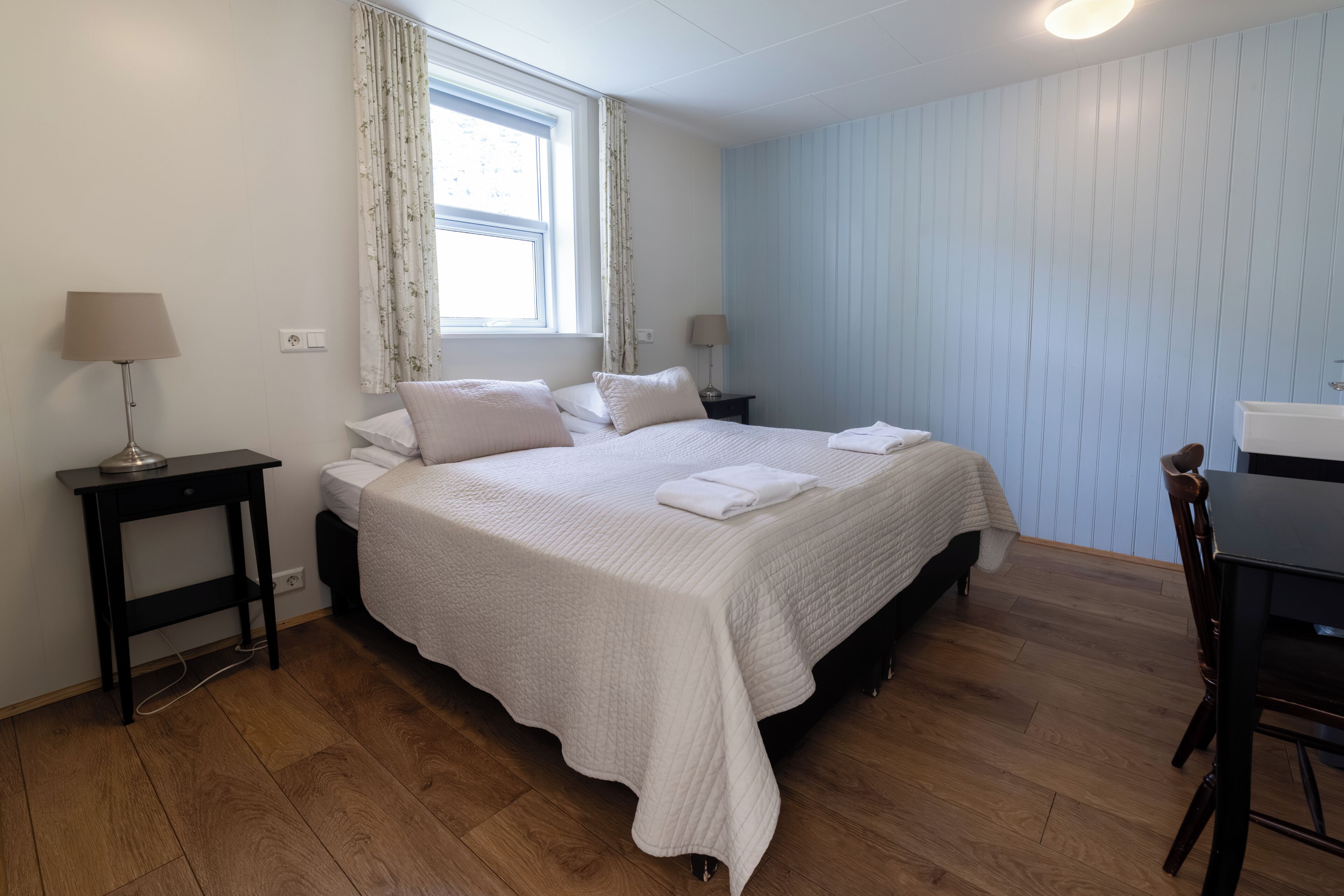 A bedroom with a bed, two bedside tables, a window with curtains, a desk, and a chair.
