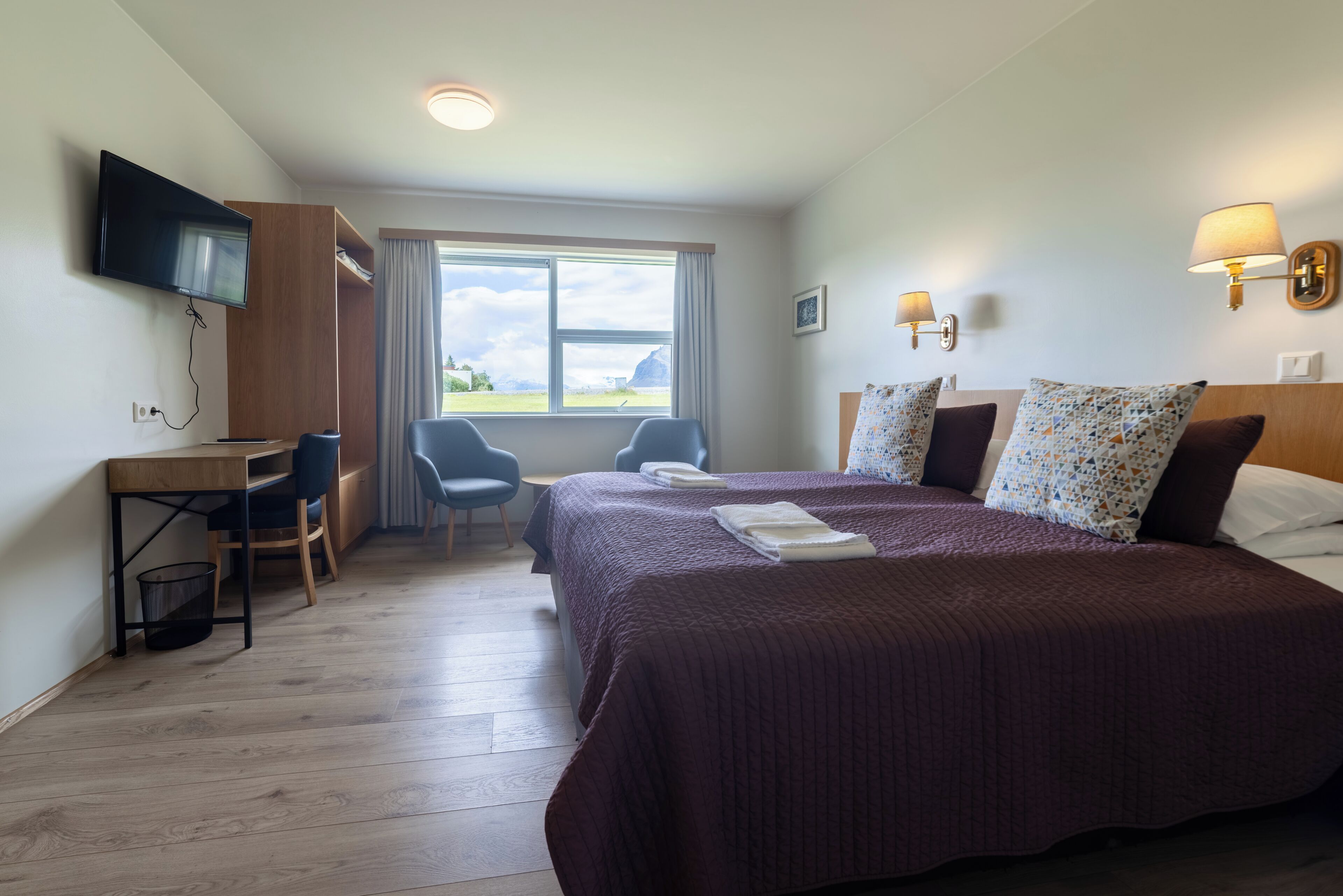 A hotel room with a bed, two bedside tables, a desk, a chair, a television, and a window with a view.