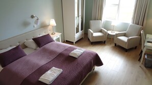 A hotel room with a large bed, two armchairs, a small table, a wardrobe, and a window with curtains.