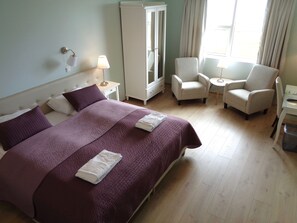 Ett hotellrum med en stor säng, två fåtöljer, ett litet bord, en garderob och ett fönster med gardiner.