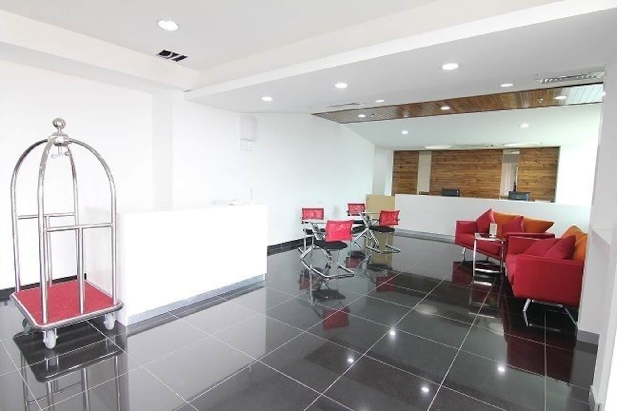 A modern hotel lobby with a reception desk, red chairs, a glass elevator, and a polished black floor.