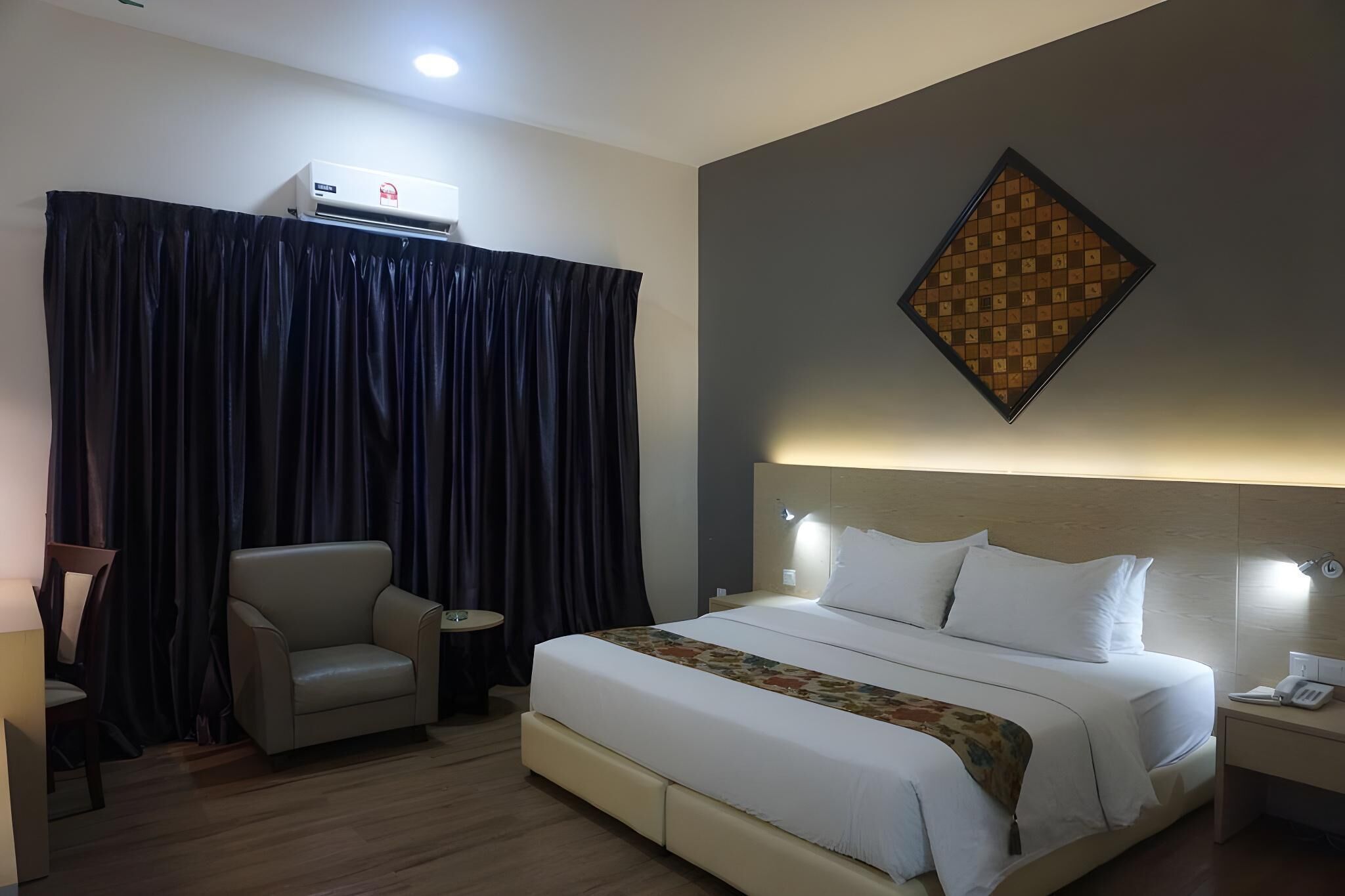A hotel room with a bed, a chair, a small table, an air conditioning unit, and a wall-mounted light fixture.