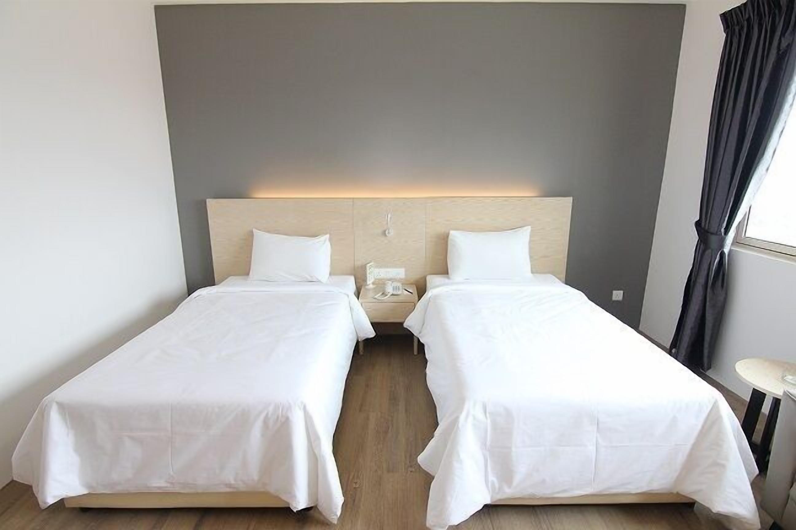 A hotel room with two single beds, a wooden headboard, a bedside table with a lamp, and a window with curtains.