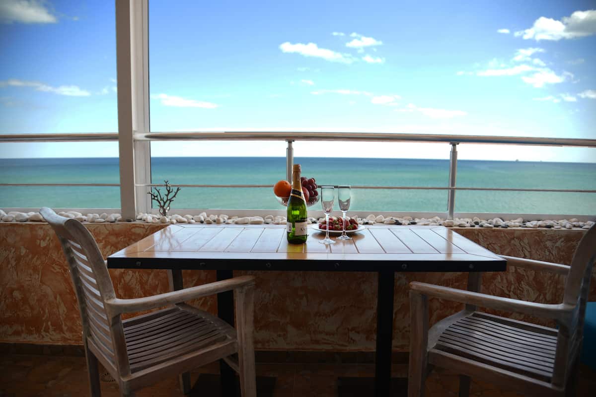 A table with a bottle of wine, two glasses, and a plate of grapes by a window overlooking the sea.