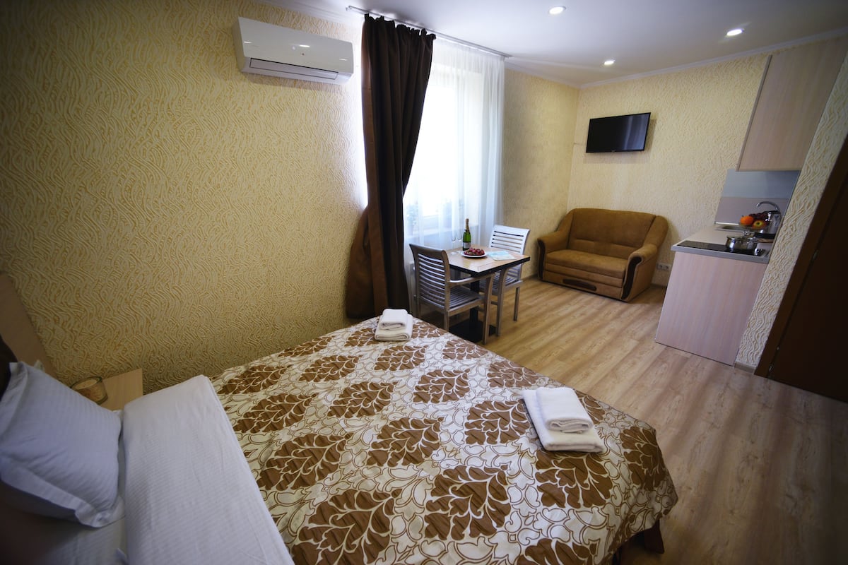 A hotel room with a bed, a chair, a small table with a bottle and a bowl of fruit, a television, and a kitchenette.