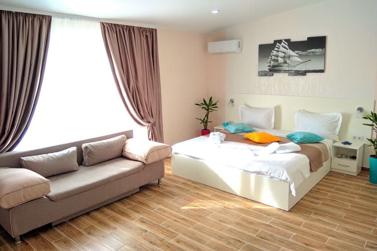 A modern hotel room with a sofa, a bed with colorful pillows, a potted plant, and a wall-mounted air conditioner.