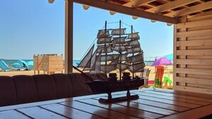 Une terrasse en bois ornée d’une maquette de bateau, d’un luminaire suspendu et offrant une vue sur la plage et les parasols.