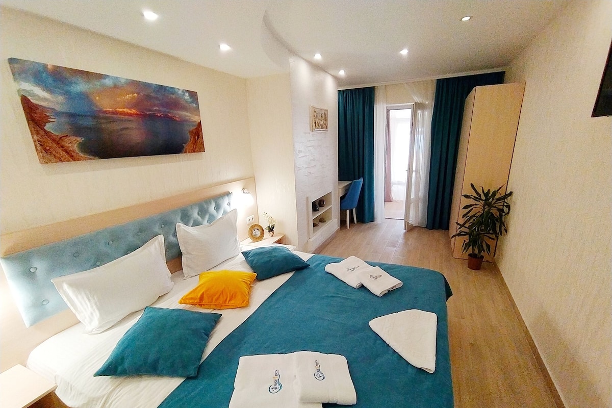 A hotel room with a large bed, a headboard, a painting, a desk, a chair, a potted plant, and a view of the outside.