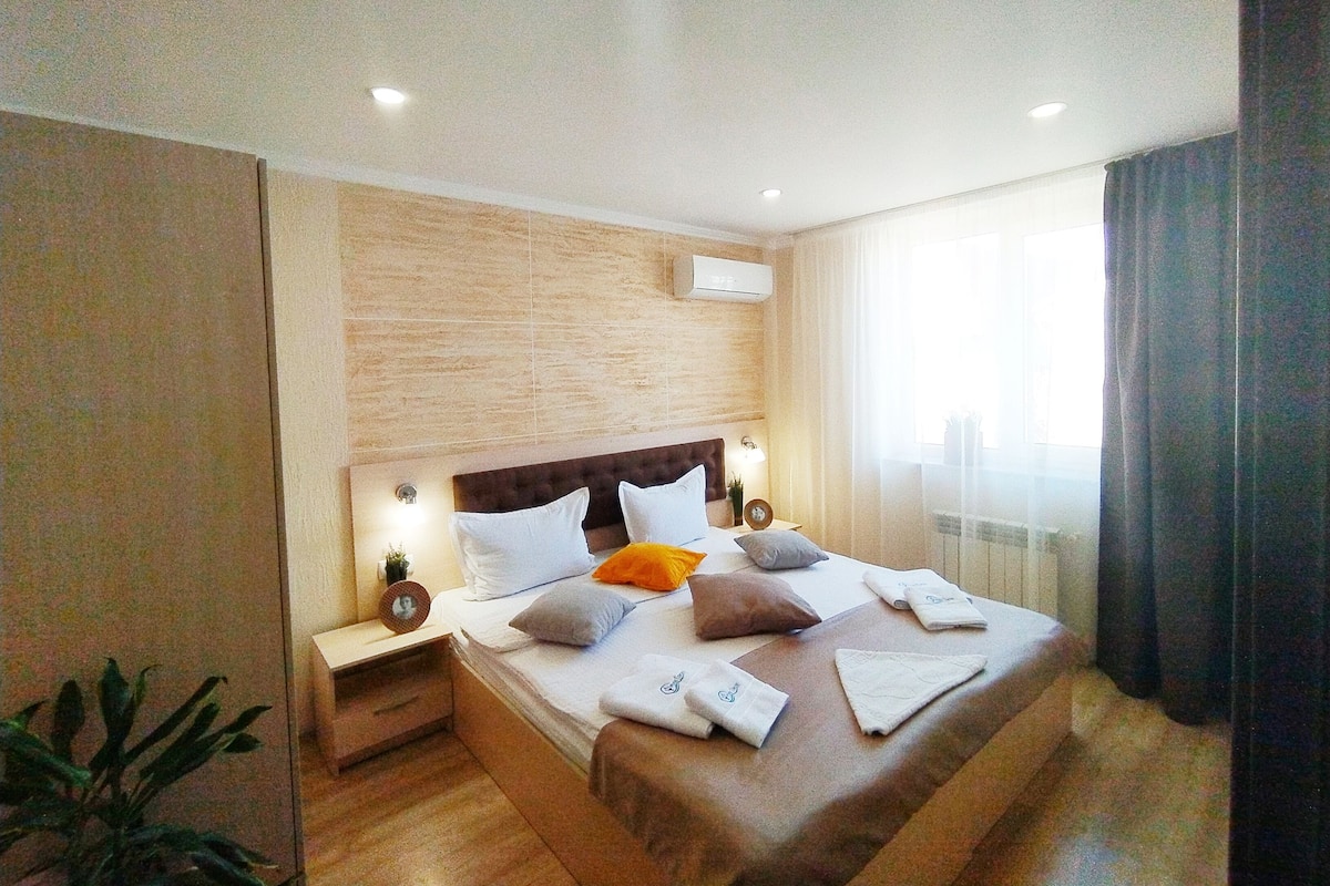 A modern hotel room with a large bed, wooden headboard, bedside tables with lamps, and a window with curtains.
