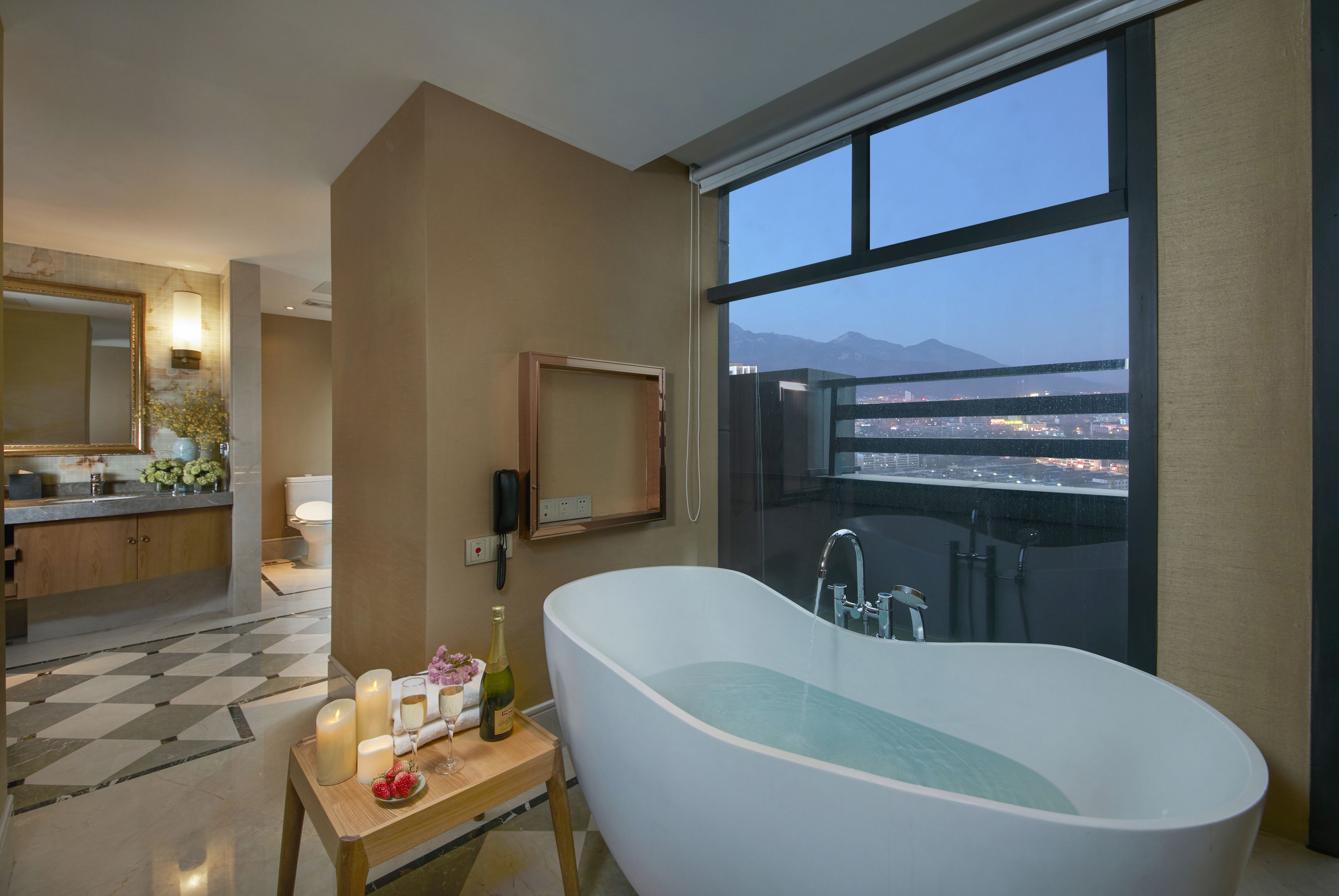 Un baño moderno con una bañera independiente, un espejo grande y vistas a un paisaje urbano a través de un ventanal amplio.