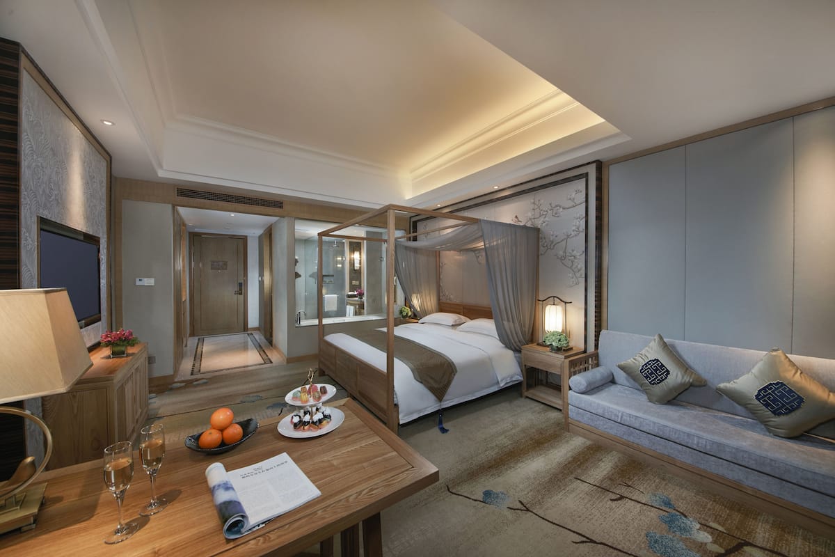 Quarto de hotel com uma cama de dossel, um sofá, uma escrivaninha com uma luminária e uma pequena mesa com laranjas e um prato de comida.