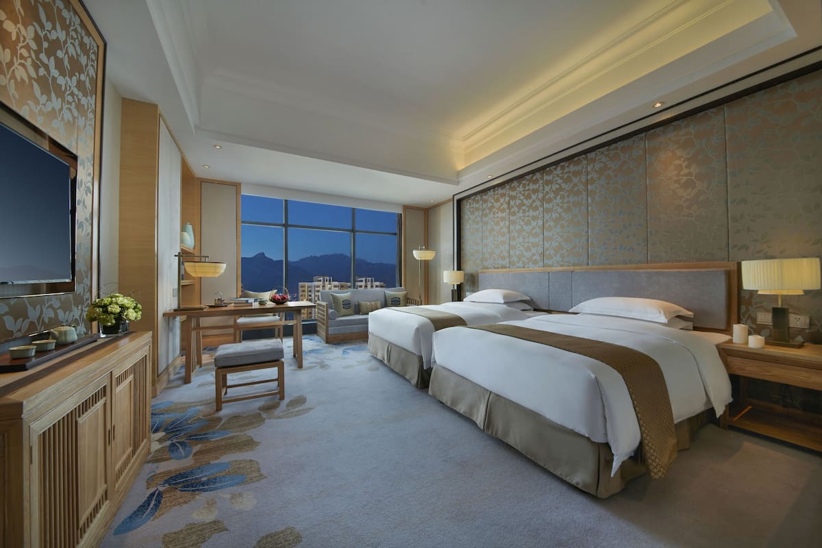 Quarto de hotel com duas camas, uma escrivaninha, uma cadeira, uma televisão e uma janela grande com vista para as montanhas.