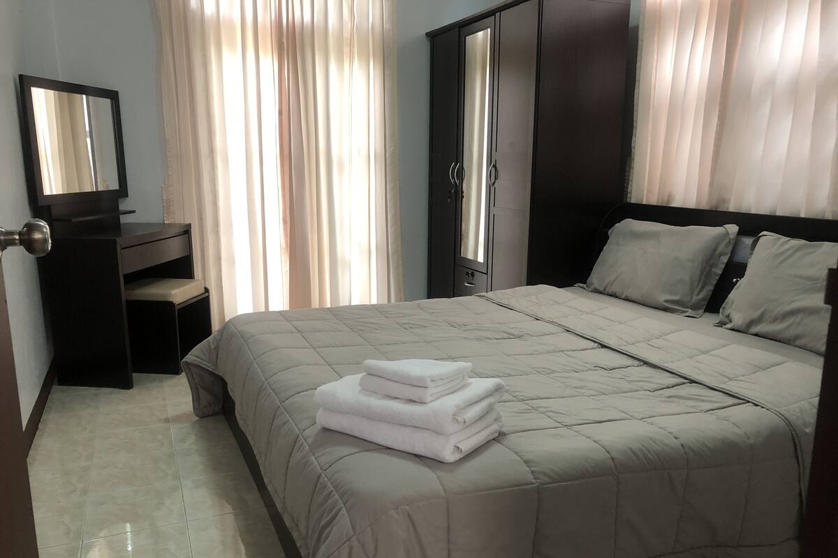 Un lit bien fait, recouvert d’une courtepointe beige, un petit bureau avec une chaise, une armoire et un miroir accroché au mur.