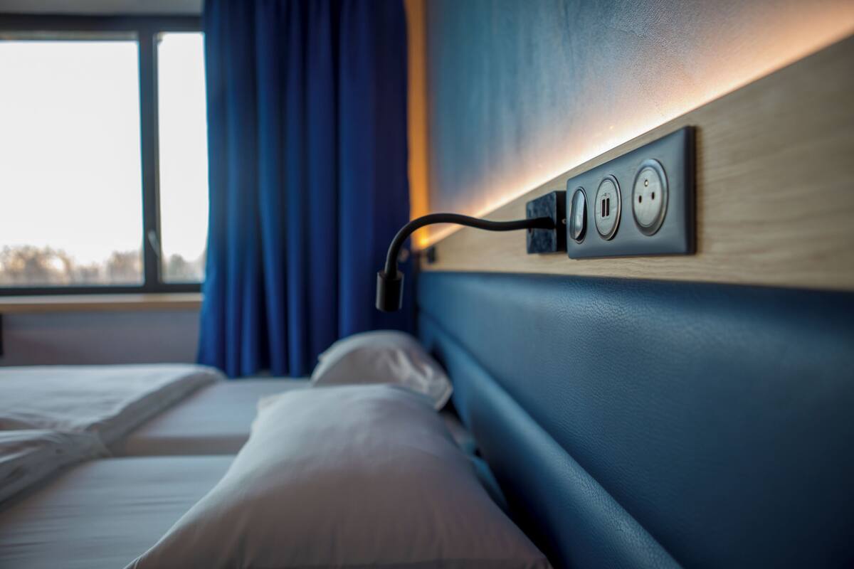 Ein Hotelzimmer mit Bett, Nachttisch und einem fest an der Wand befestigten Steckdosenleisten mit drei Steckern.
