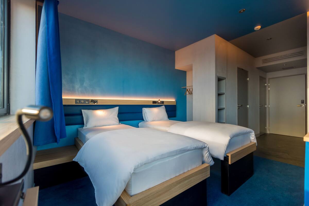 Ein modernes Hotelzimmer mit zwei Betten, einer blauen Akzentwand und einem hölzernen Kopfteil.