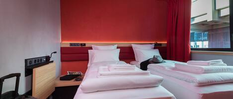 Habitación de hotel con dos camas, un escritorio, una silla y un ventanal con cortinas rojas.