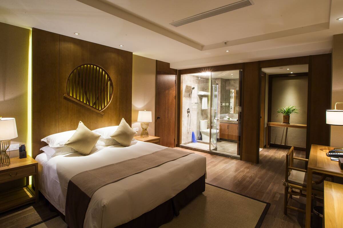 A hotel room with a large bed, bedside lamps, a desk with a chair, a bathroom with a shower, and a potted plant.