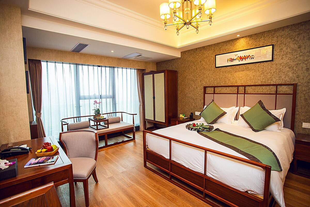 A hotel room with a large bed, a desk with a chair, a small table with a chair, and a large window with curtains.