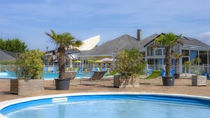 A swimming pool with lounge chairs and palm trees, surrounded by a fenced area with a building in the background. - Village de vacances Le Sénéquet (Blainville-sur-Mer)