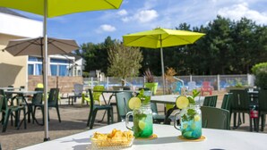 An outdoor dining area with tables and chairs, two drinks with lime and mint, and a bright yellow umbrella. - Village de vacances Le Sénéquet (Blainville-sur-Mer)