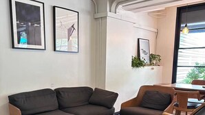 A modern living room with a sofa, two armchairs, and a small wooden table. There are two framed pictures on the wall, a potted plant, and a window with a view. - T Hostel at Victory Monument (Bangkok)