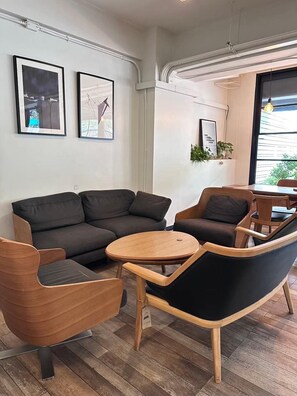 Una sala moderna con un sofá, dos sillones y una pequeña mesa de madera. Hay dos cuadros en la pared, una planta en maceta y una ventana con vistas.