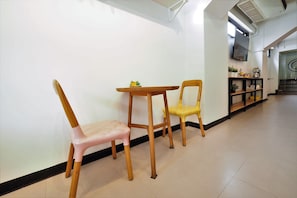 Un pasillo con dos sillas de madera y una mesita, un estante negro con plantas, y una escalera al fondo.