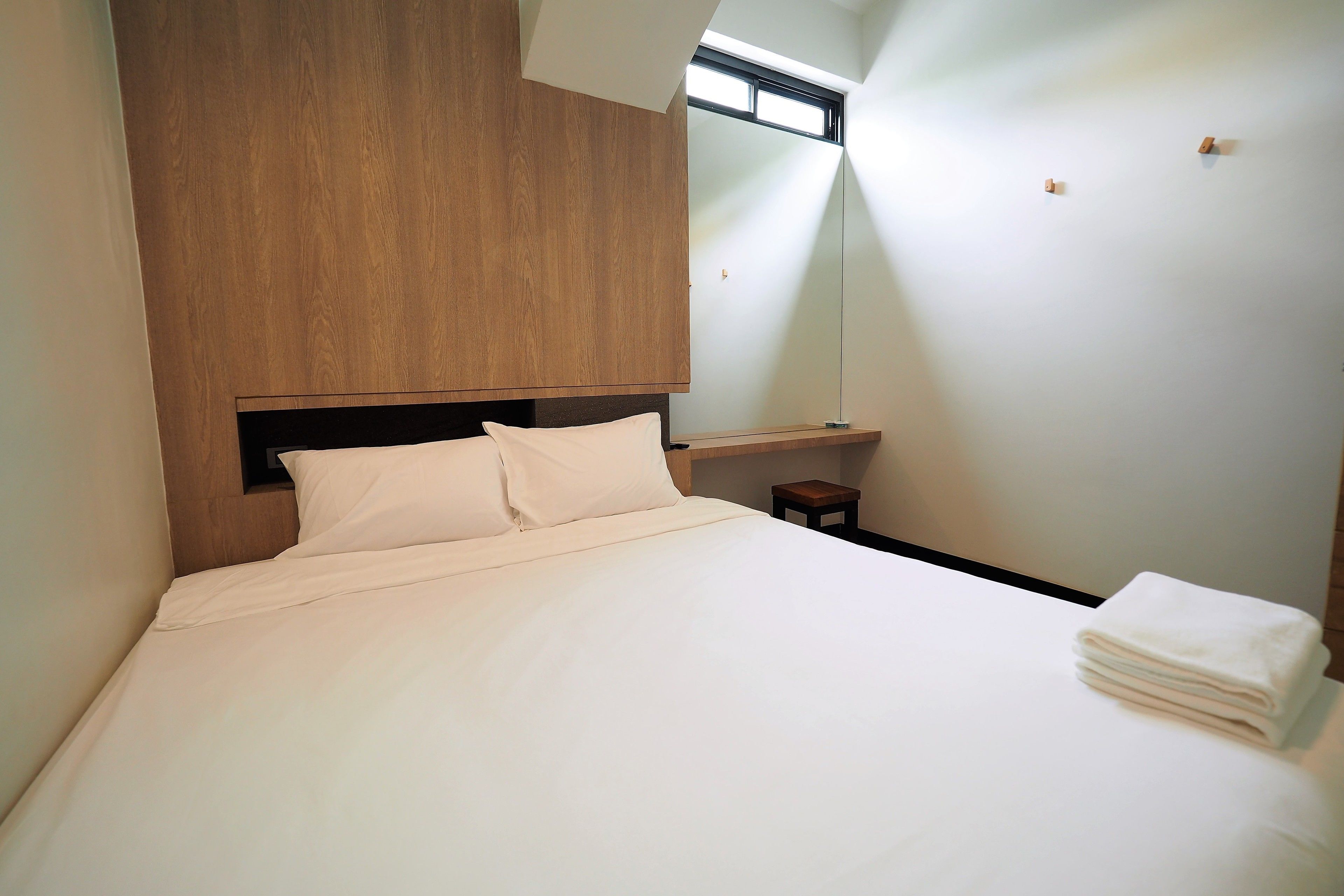 A neatly made bed with white linens, a wooden headboard, a small bedside table with a folded white towel, and a large mirror on the wall.