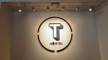 Un logotipo circular con una 'T' estilizada y la palabra 'HOSTEL' debajo, iluminado por focos.