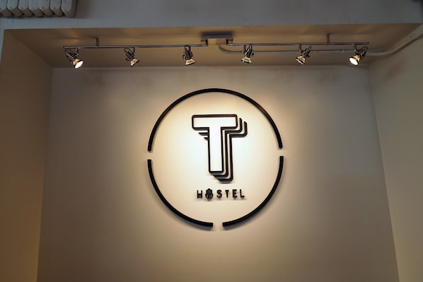 A circular logo with a stylized 'T' and the word 'HOSTEL' below it, illuminated by spotlights. - T Hostel at Victory Monument (Bangkok)