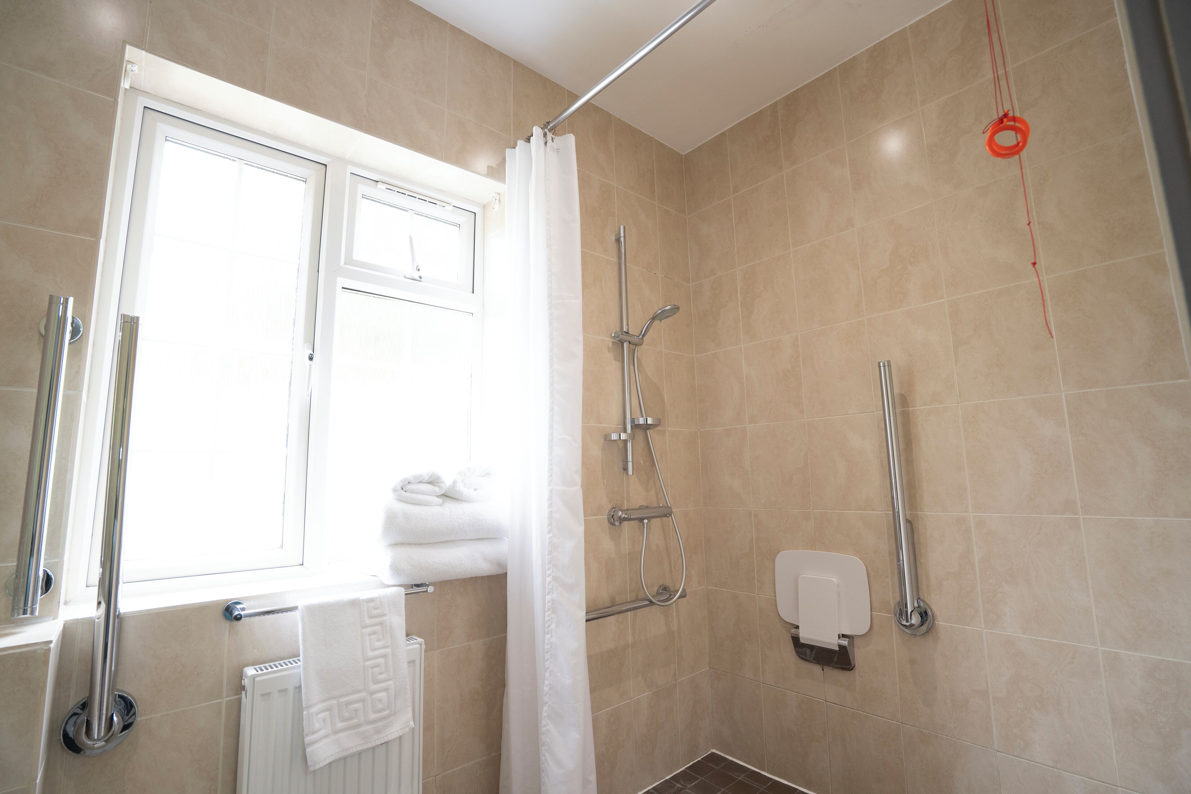A bathroom with a shower, a toilet, a handrail, and a window with a curtain.