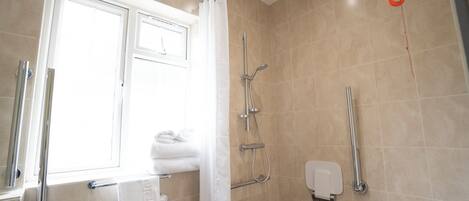 A bathroom with a shower, a toilet, a handrail, and a window with a curtain.
