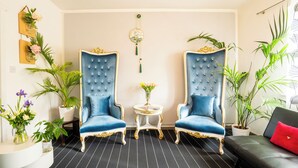 Een kamer met twee blauwe fauteuils, een klein rond tafeltje met een bloevas en een zwarte bank met een rode kussens.