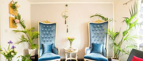 A room with two blue upholstered armchairs, a small round table with a vase of flowers, and a black sofa with a red cushion.