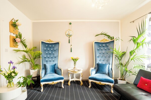Una habitación con dos sillones tapizados en azul, una mesita redonda con un florero y un sofá negro con un cojín rojo.