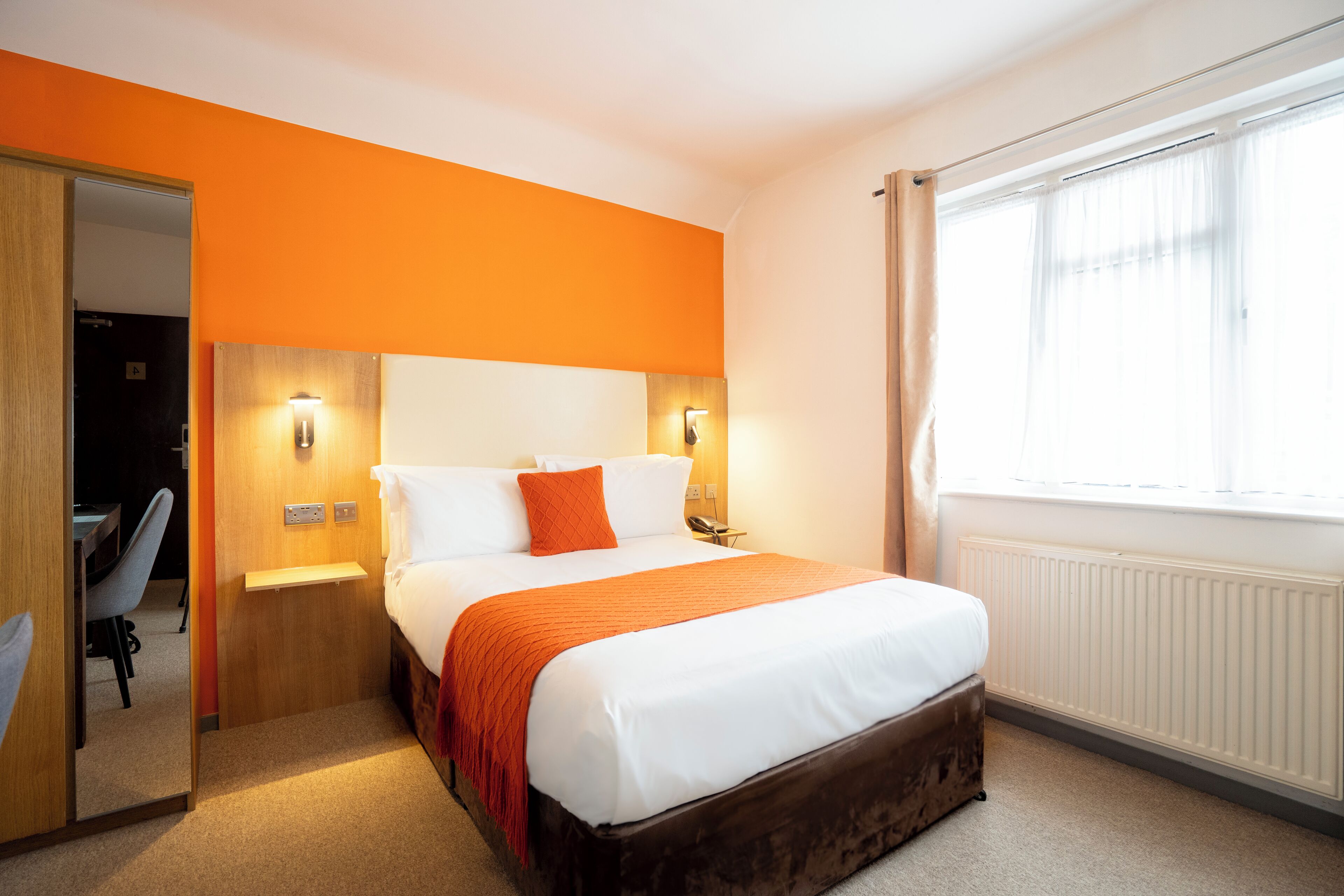 A hotel room with a large bed, a wooden headboard, two bedside lamps, a window with curtains, and a door leading to a bathroom.