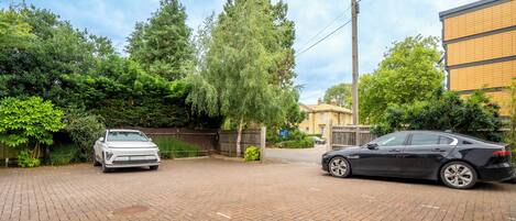 A paved driveway with two parked cars, surrounded by trees and a building with a yellow facade.