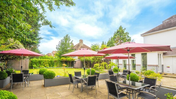 Een terras buiten met tafels en stoelen, rode parasols en potplanten.